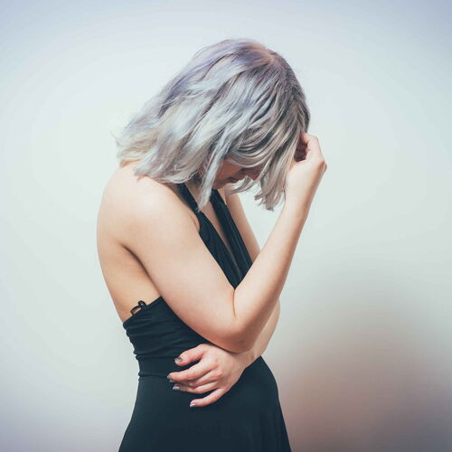 Eine Frau steht mit gesenktem Kopf. Ihr Gesicht ist von den Haaren verdeckt. Sie hält sich die Hand an den Kopf und wirkt bedrückt.